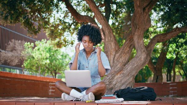ラップトップと緑の木の下に座っているヘッドフォンを着ているティーンガール. 美しいアフリカ系アメリカ人のティーンエイジャーは,自然に関するオンライン研究のためにイヤホンを置きます. コンピュータ上のスマートな女性学生のリスニングコース. - 写真・画像