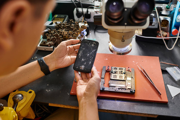 καλλιεργημένη άποψη πεπειραμένου τεχνικού που κρατά κινητό τηλέφωνο με σπασμένη οθόνη κοντά σε εξοπλισμό επισκευής - Φωτογραφία, εικόνα