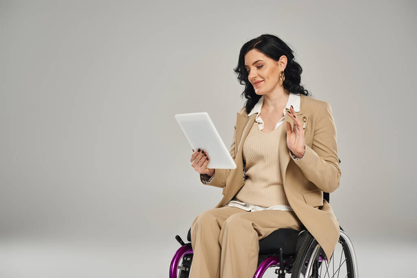 cheerful beautiful disabled woman in pastel attire in wheelchair waving at tablet during video call - Photo, Image