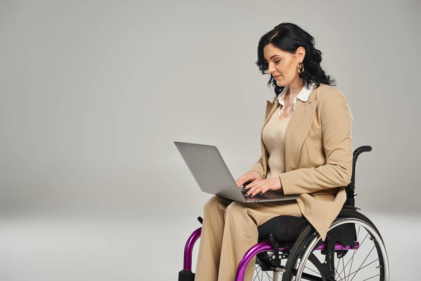 hermosa mujer discapacitada con confianza en silla de ruedas con traje de pastel y trabajando en su computadora portátil - Foto, Imagen