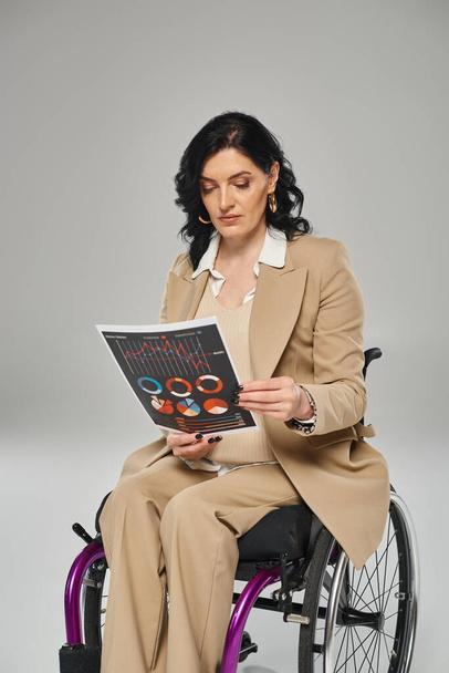 brunette vrouw met mobiliteit handicap in pastel pak kijken naar graphics terwijl in een rolstoel - Foto, afbeelding