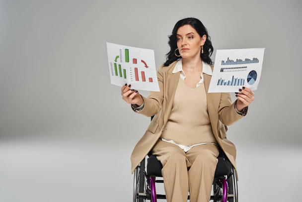séduisante femme handicapée avec les cheveux ondulés en costume chic en fauteuil roulant en regardant les graphiques dans ses mains - Photo, image