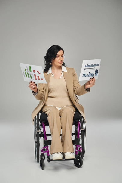 pewna siebie kobieta z niepełnosprawnością ruchową w eleganckim garniturze na wózku inwalidzkim patrząc na jej grafikę - Zdjęcie, obraz