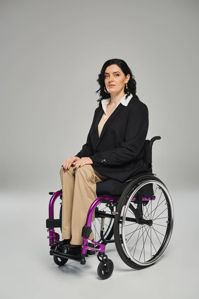 attraktive Frau mit Behinderung im schwarzen Blazer im Rollstuhl sitzend und in die Kamera blickend - Foto, Bild
