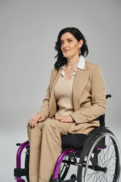 selbstbewusste brünette Frau mit Behinderung im pastellfarbenen Outfit im Rollstuhl sitzend und wegschauend - Foto, Bild
