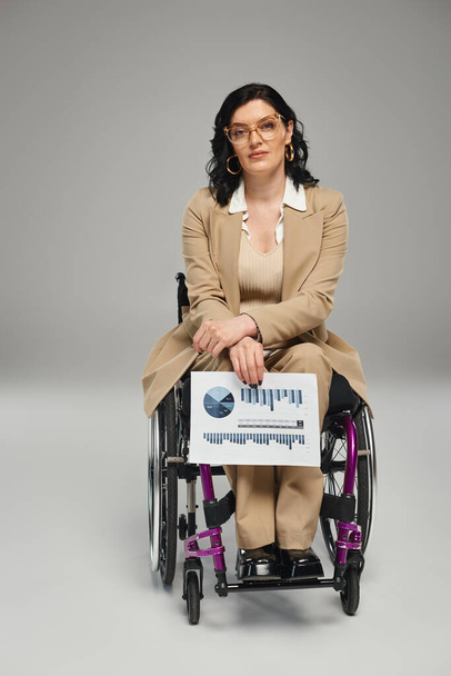 atrakcyjna niepełnosprawna kobieta w okularach na wózku inwalidzkim patrząca w kamerę z grafiką w ręku - Zdjęcie, obraz