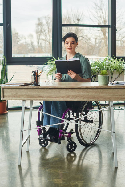 boa aparência confiante mulher de negócios com deficiência em cadeira de rodas olhando para seu caderno no escritório - Foto, Imagem