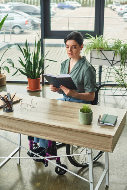 femme brune confiante en tenue décontractée en fauteuil roulant travaillant avec son ordinateur portable au bureau - Photo, image