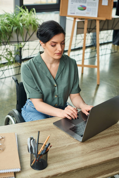 mulher de negócios de boa aparência em traje casual em cadeira de rodas trabalhando em seu laptop enquanto no escritório - Foto, Imagem
