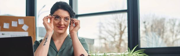 mulher deficiente atraente em roupa casual com tatuagem e óculos olhando para longe, enquanto no escritório - Foto, Imagem