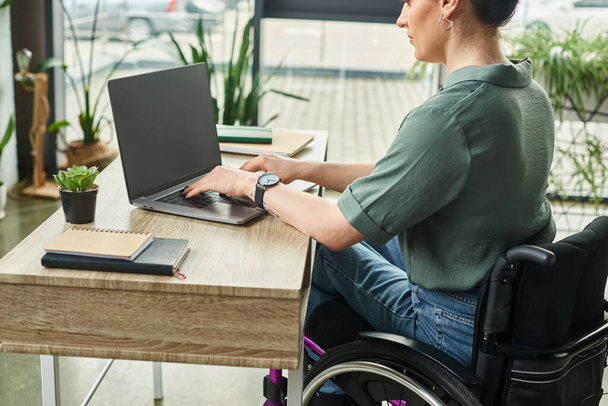 ελκυστική επιχειρηματίας με αναπηρία με περιστασιακή ενδυμασία σε αναπηρικό αμαξίδιο που κάθεται στο φορητό υπολογιστή που εργάζεται στο γραφείο - Φωτογραφία, εικόνα