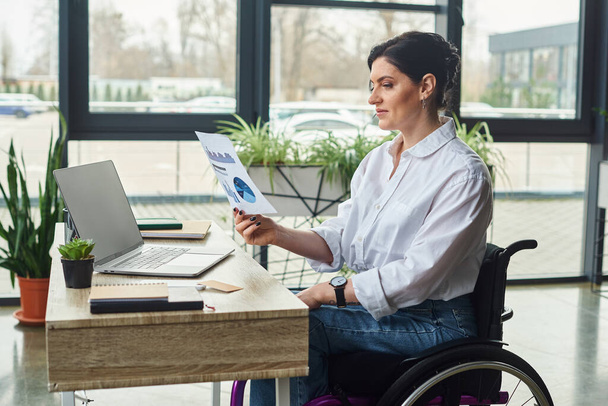 dedykowane bizneswoman z niepełnosprawnością na wózku inwalidzkim patrząc na jej grafiki w biurze - Zdjęcie, obraz