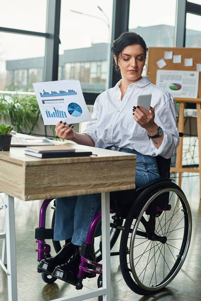 mulher deficiente atraente em traje elegante em cadeira de rodas olhando para o telefone enquanto trabalhava com gráficos - Foto, Imagem