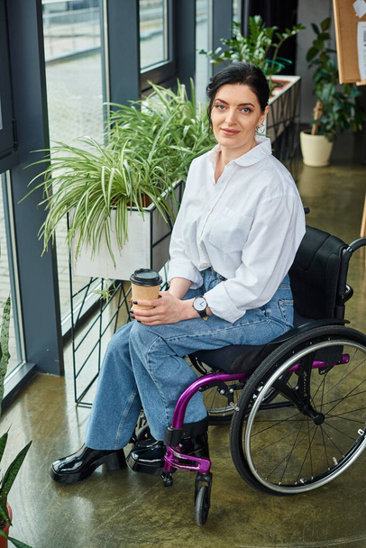 veselá zdravotně postižená podnikatelka ve stylovém oblečení v invalidním vozíku drží kávu a dívá se do kamery - Fotografie, Obrázek