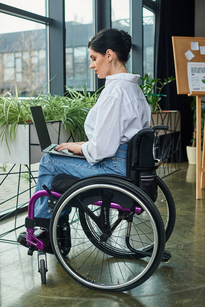 toegewijde zakenvrouw met een handicap in stedelijke kleding in rolstoel werken op haar laptop in kantoor - Foto, afbeelding