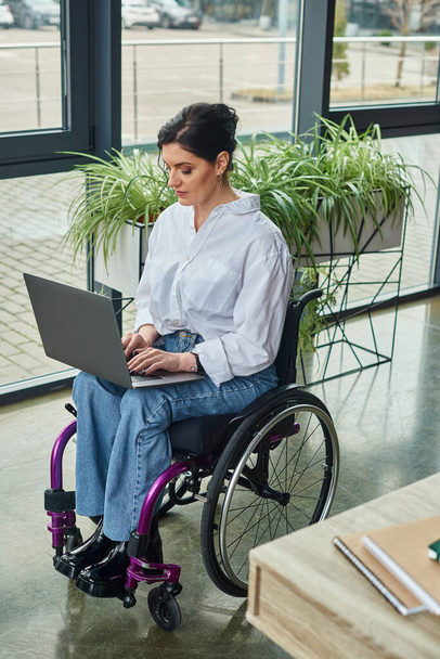 Görevdeyken laptopta çalışan, tekerlekli sandalyeli, sıradan giyimli, engelli bir iş kadını. - Fotoğraf, Görsel