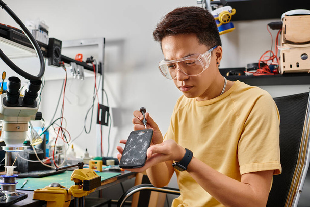 asiatischer Techniker mit Brille öffnet Smartphone mit kaputtem Bildschirm mit Schraubenzieher in Werkstatt - Foto, Bild