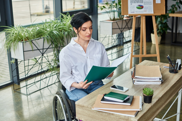 confident disabled businesswoman in wheelchair looking at her paperwork at her workplace in office - Photo, Image