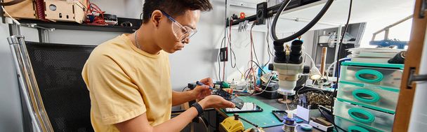 Gözlüklü yetenekli Asyalı teknisyen tamir atölyesi ve afişte elektronik aletlerle çalışıyor. - Fotoğraf, Görsel