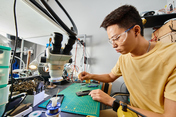 концентрированный азиатский ремонтник в очках делает техническую экспертизу мобильного телефона в ремонтной мастерской - Фото, изображение