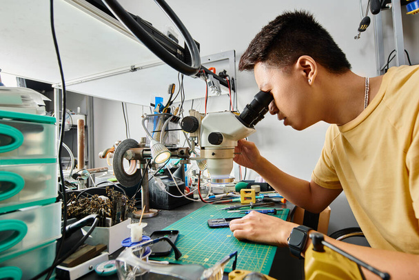side view of skilled asian technician looking in microscope while working with electronic equipment - Photo, Image
