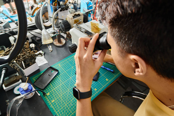 Hochwinkelaufnahme des gelernten Reparateurs bei der Arbeit mit Mikroskop und elektronischen Geräten in der Werkstatt - Foto, Bild