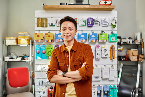 szczęśliwy azjatycki kierownik sklepu uśmiechnięty do kamery w pobliżu licznika prywatnego sklepu elektronicznego, mały biznes - Zdjęcie, obraz