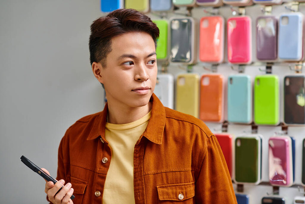 серьезный азиат держит мобильный телефон и смотрит в сторону в частном магазине электроники, малый бизнес - Фото, изображение