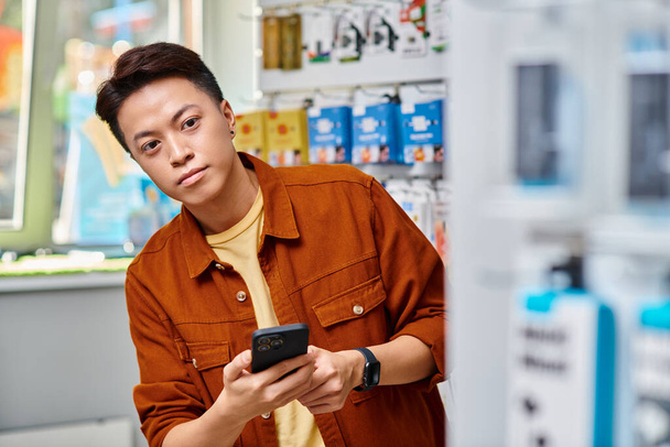 asijský podnikatel držící mobilní telefon a dívající se pryč ve vlastním obchodě s elektronikou, malé podnikání - Fotografie, Obrázek