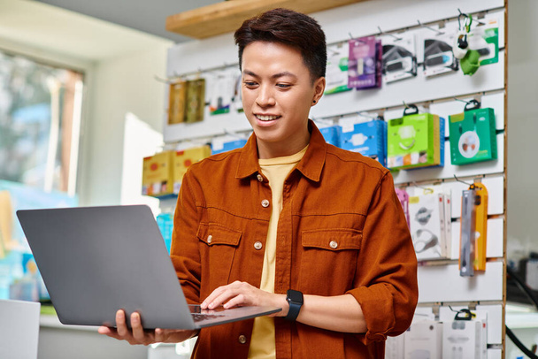 felice imprenditore asiatico utilizzando computer portatile in piedi nel proprio negozio di elettronica, concetto di piccola impresa - Foto, immagini