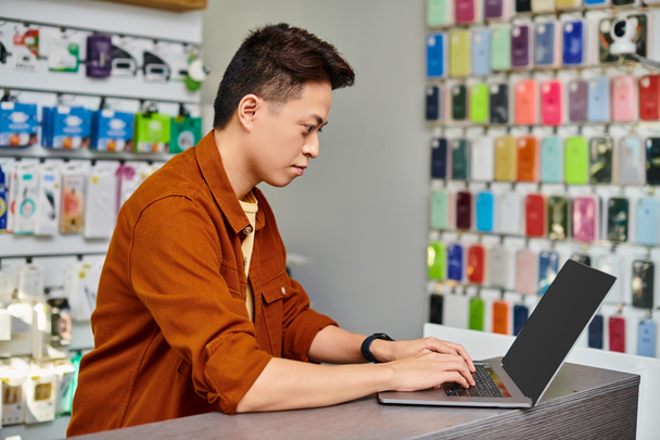 συγκεντρωμένος ασιατικός άνθρωπος που χρησιμοποιεί το lap-top στον πάγκο στο ιδιωτικό κατάστημα ηλεκτρονικών, έννοια των μικρών επιχειρήσεων - Φωτογραφία, εικόνα