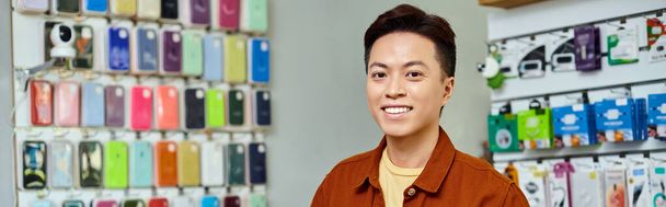 glimlachende succesvolle Aziatische ondernemer op zoek naar camera in particuliere elektronica winkel, banner - Foto, afbeelding