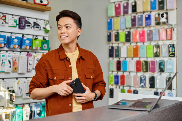 feliz asiático homem de negócios com telefone móvel olhando para longe perto de laptop na loja de eletrônicos privada - Foto, Imagem