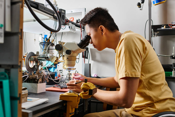 Seitenansicht des professionellen Asienspezialisten mit Mikroskop, der das elektronische Gerät in der Werkstatt untersucht - Foto, Bild