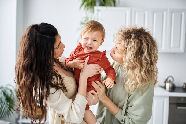 liefhebbende lgbt paar in homewear houden hun baby meisje in handen en troosten haar, familie concept - Foto, afbeelding