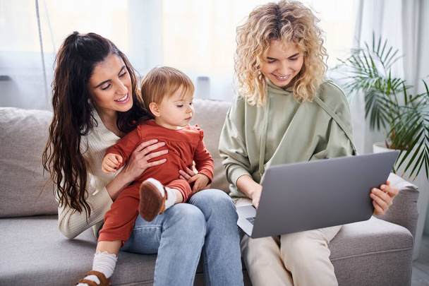 feliz pareja de lesbianas amorosas sentados en el sofá con su niña mirando a la computadora portátil, concepto de familia - Foto, Imagen