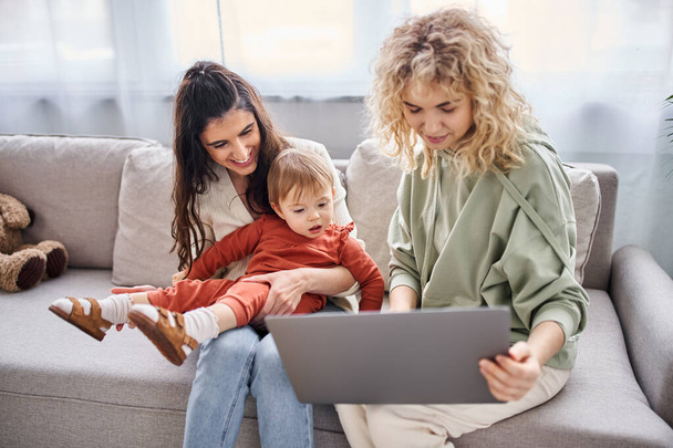 fröhlich liebendes lesbisches Paar auf Sofa sitzend mit ihrem kleinen Mädchen, das auf Laptop schaut, Familienkonzept - Foto, Bild