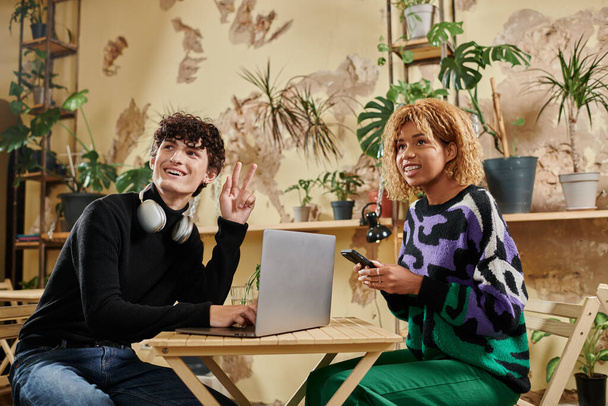 homme heureux appelant un serveur tout en utilisant un ordinateur portable et assis avec une fille afro-américaine dans un café végétalien - Photo, image
