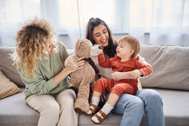attractive jolly lesbian couple with their toddler daughter and teddy bear on sofa, family concept - Fotó, kép