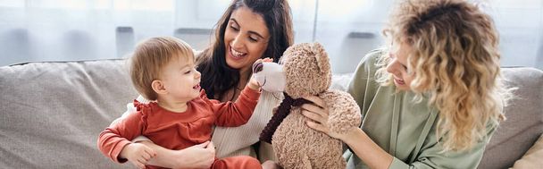 jolly lesbian couple with their toddler daughter and teddy bear on sofa, family concept, banner - Photo, Image