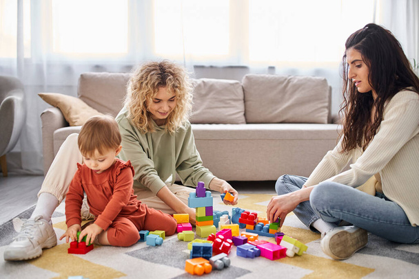 cheerful appealing lesbian couple playing with their toddler daughter at home, modern parenting - Photo, Image