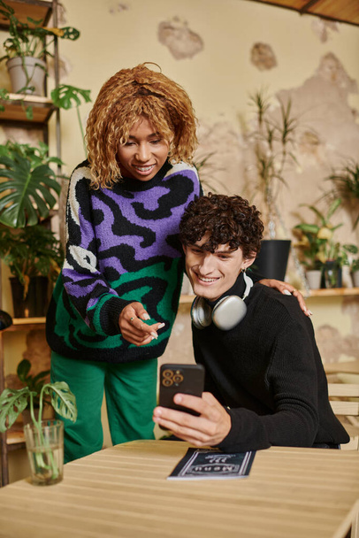 happy multicultural couple with curly hair looking at smartphone and choosing menu in vegan cafe - Photo, Image