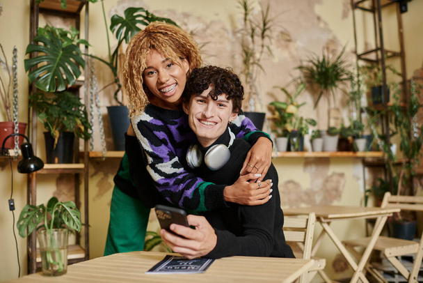 cheerful african american woman with curly hair embracing young man with  smartphone in vegan cafe - Foto, afbeelding