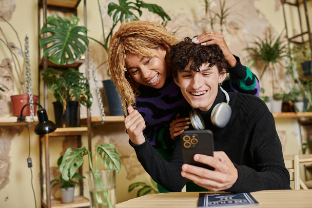 jolly donna afroamericana con i capelli ricci abbracciando giovane uomo con smartphone in caffè vegan - Foto, immagini