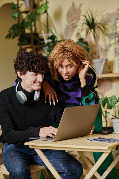 heureuse femme afro-américaine avec bretelles debout près du petit ami et regardant ordinateur portable dans le café - Photo, image