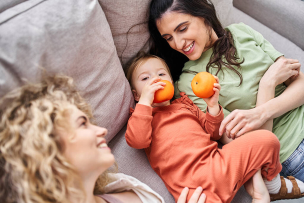 fröhliches lgbt-Paar auf dem Sofa liegend mit ihrem hübschen kleinen Mädchen, das Mandarinen hält, Familienkonzept - Foto, Bild