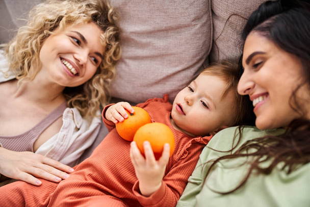 merry lgbt couple lying on sofa with their pretty baby girl holding tangerines, family concept - Photo, Image