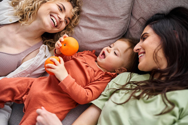 beautiful happy lgbt couple lying on sofa with their baby girl holding tangerines, family concept - Photo, Image