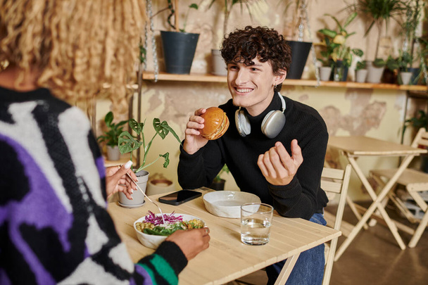 happy young man in headphones holding tofu burger and looking at african american woman in cafe - Photo, Image