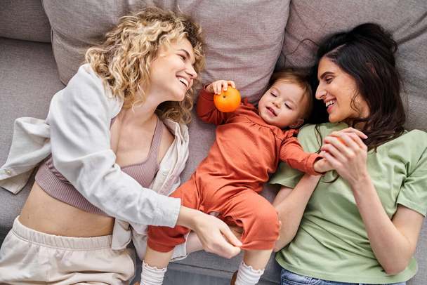 beautiful joyous lgbt couple lying on sofa with their baby girl holding tangerines, family concept - Photo, Image